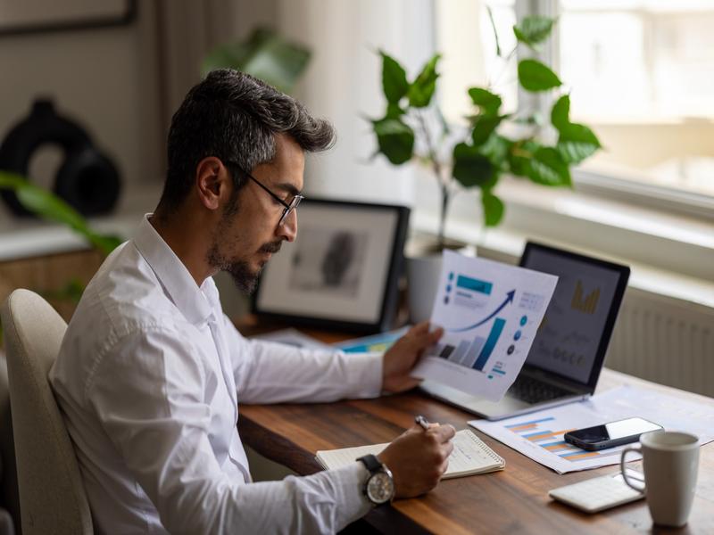 Homme travaillant depuis son bureau, analysant des graphiques financiers imprimés et prenant des notes.