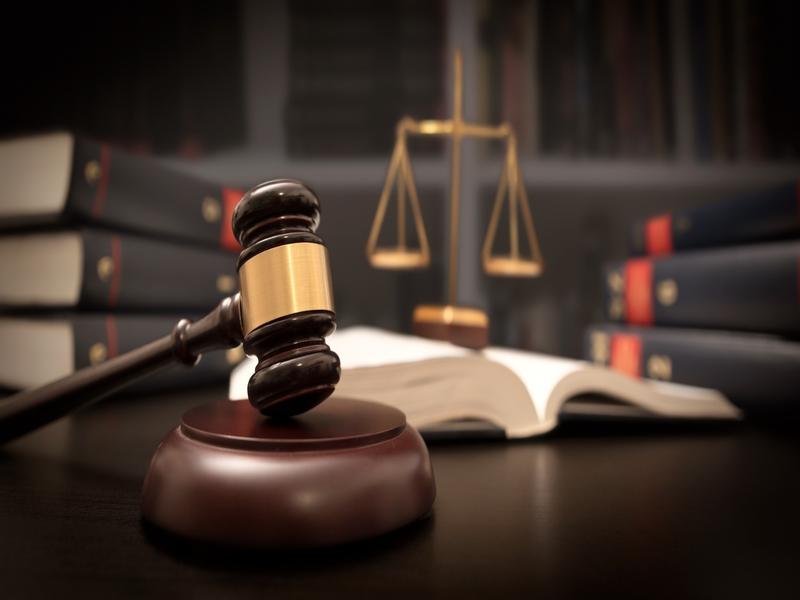 Marteau de juge posé sur son socle, avec en arrière-plan un livre ouvert et une balance dorée, symbole de justice.