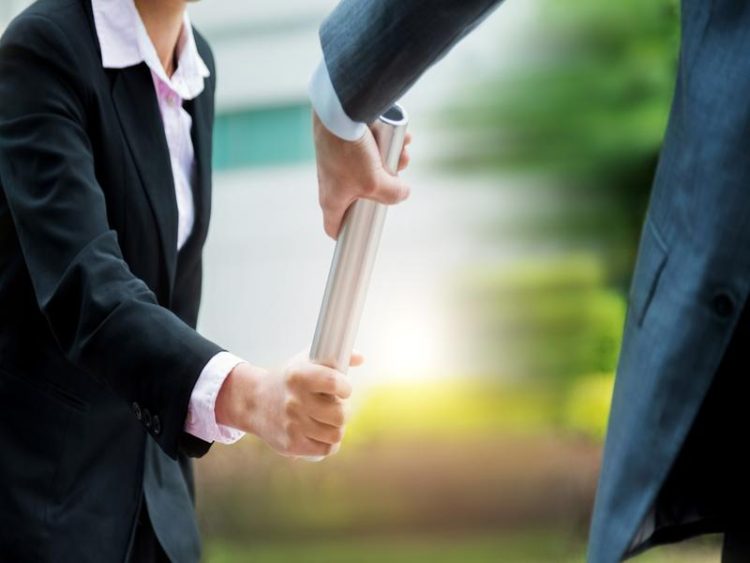 Gros plan sur deux personnes en costume se passant un témoin argenté, symbolisant la relève et le transfert de responsabilités.