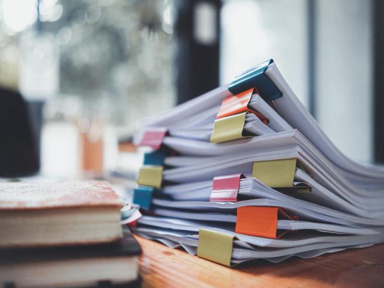 Pile de documents imprimés avec de grosses pinces colorées (jaune, bleu, rouge, orange) posés sur un bureau en bois. À gauche, un carnet est visible, ambiance bureau lumineux flou en arrière-plan.