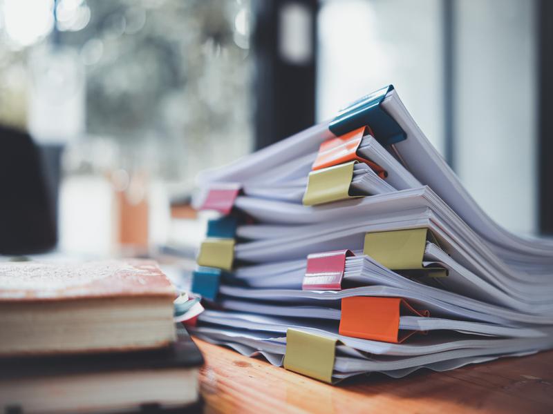 Pile de documents imprimés avec de grosses pinces colorées (jaune, bleu, rouge, orange) posés sur un bureau en bois. À gauche, un carnet est visible, ambiance bureau lumineux flou en arrière-plan.