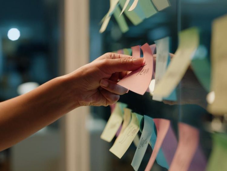 Gros plan d’une main collant un post-it coloré sur une vitre remplie de notes adhésives. Ambiance de brainstorming en équipe.