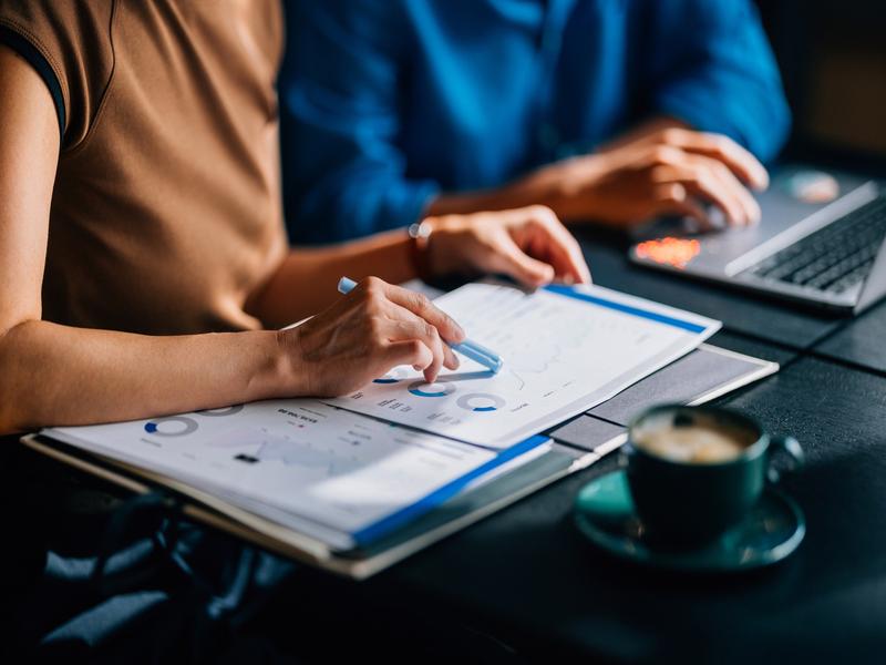 Gros plan d’une main féminine tenant un stylo bleu et pointant des graphiques imprimés sur papier. Ordinateur portable en arrière-plan, ambiance café et réunion.