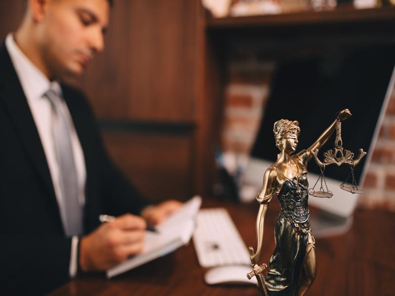 Statue de la déesse Thémis tenant la balance de la justice, devant un homme en costume écrivant.
