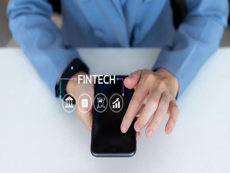 Gros plan sur une personne en veste bleue utilisant un smartphone. Le mot FINTECH apparaît en surimpression, accompagné d’icônes symbolisant la banque, les taux, l’intelligence artificielle et la croissance.