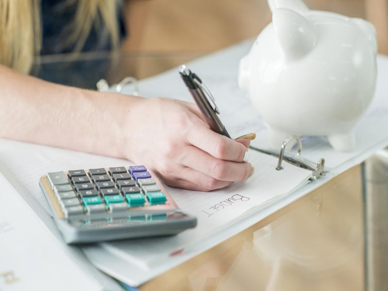 Main écrivant dans un cahier « Budget » avec une tirelire et une calculatrice.