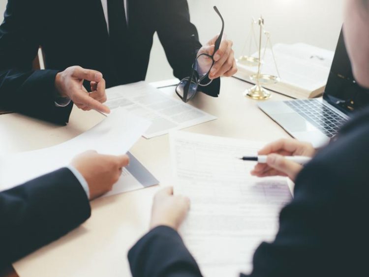 Réunion de travail entre personnes en costume, discutant et échangeant des documents juridiques, avec une balance dorée et un ordinateur portable sur la table.